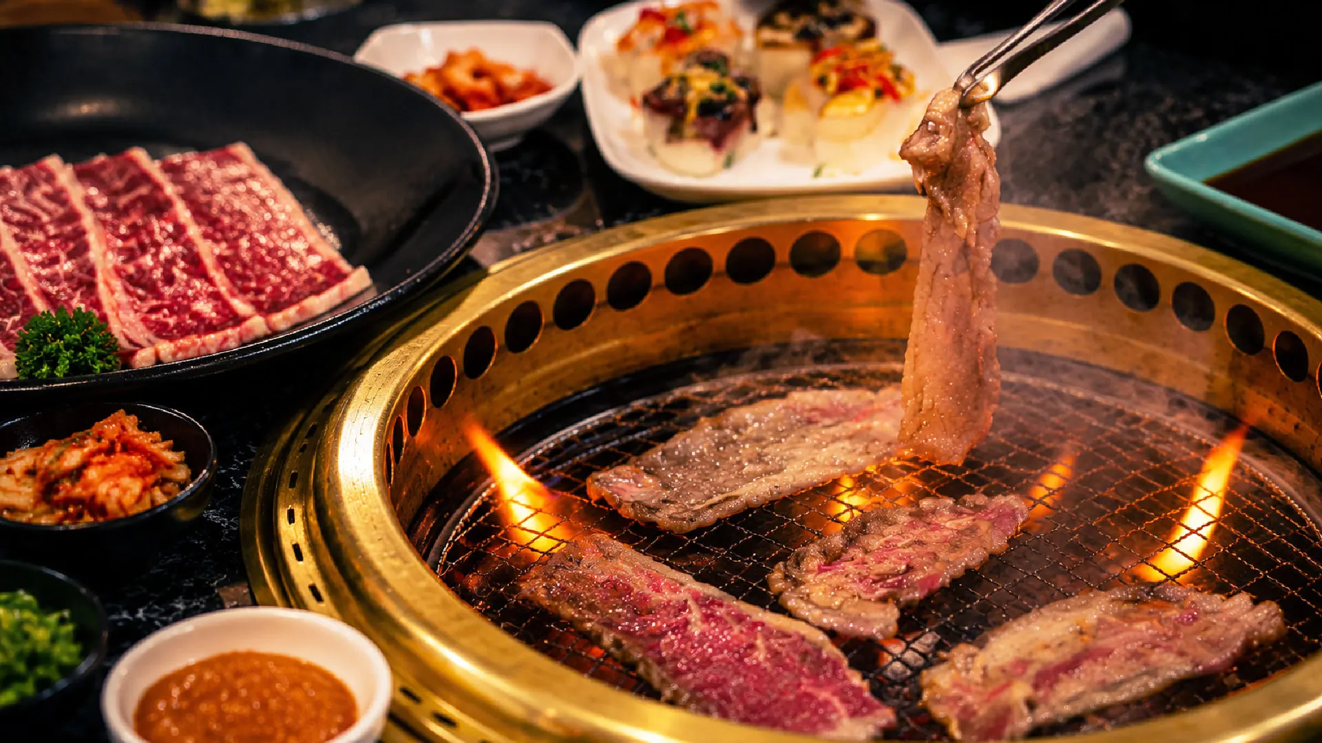 Marbled beef searing on a Gyumono tabletop grill, with sushi and side dishes around it