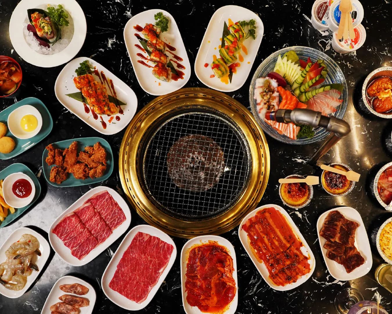 Overhead view of a full Gyumono table: marbled beef, sushi rolls, sashimi and sides around a lit grill
